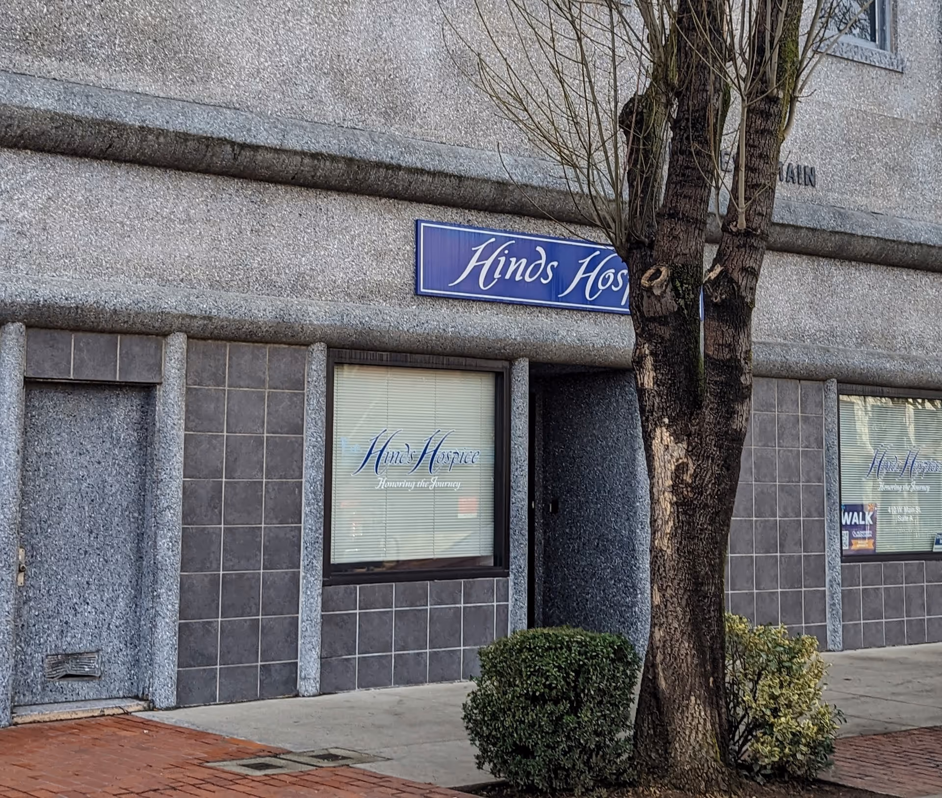 Exterior view of a grey stone building with a blue Hinds LifeCare sign and tree nearby.
