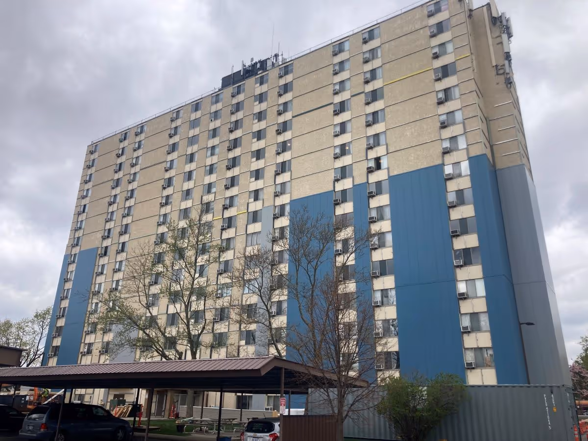 Tall beige and blue high-rise senior living building with a covered entrance and trees.