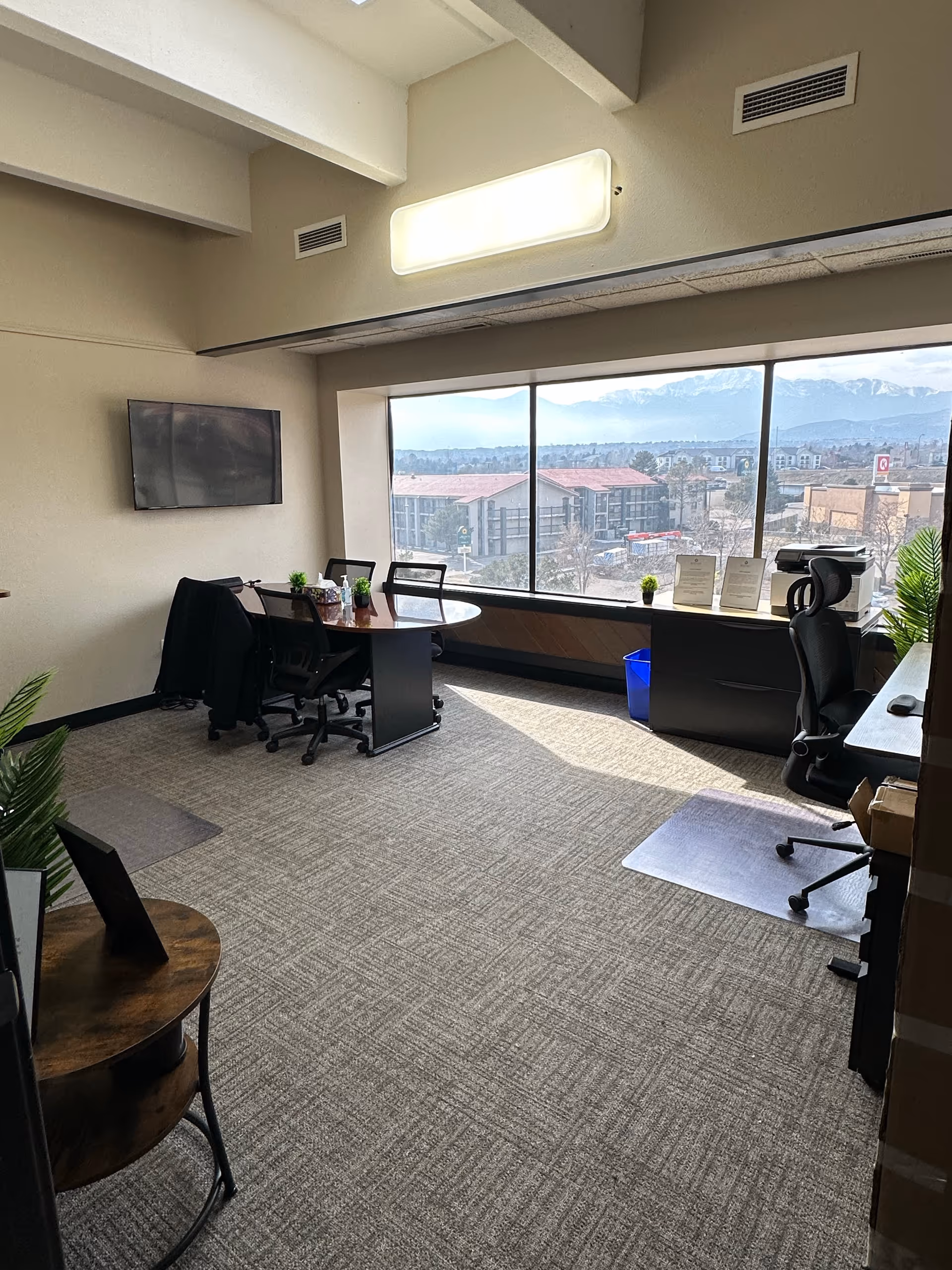 An office-like common area with desks, chairs, a wall-mounted TV, and a large window view.