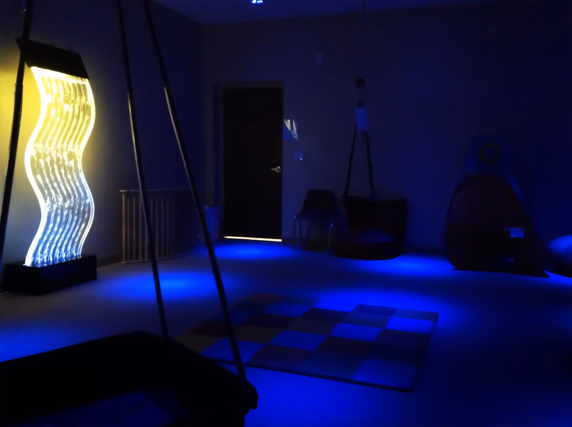 Dimly lit therapy room with blue lighting, exercise equipment, and a glowing wavy sculpture.