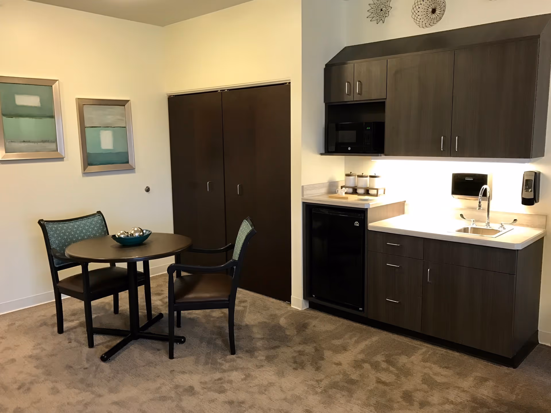Small studio kitchenette with dark cabinets, a sink, microwave and mini-fridge next to a round table and two chairs.