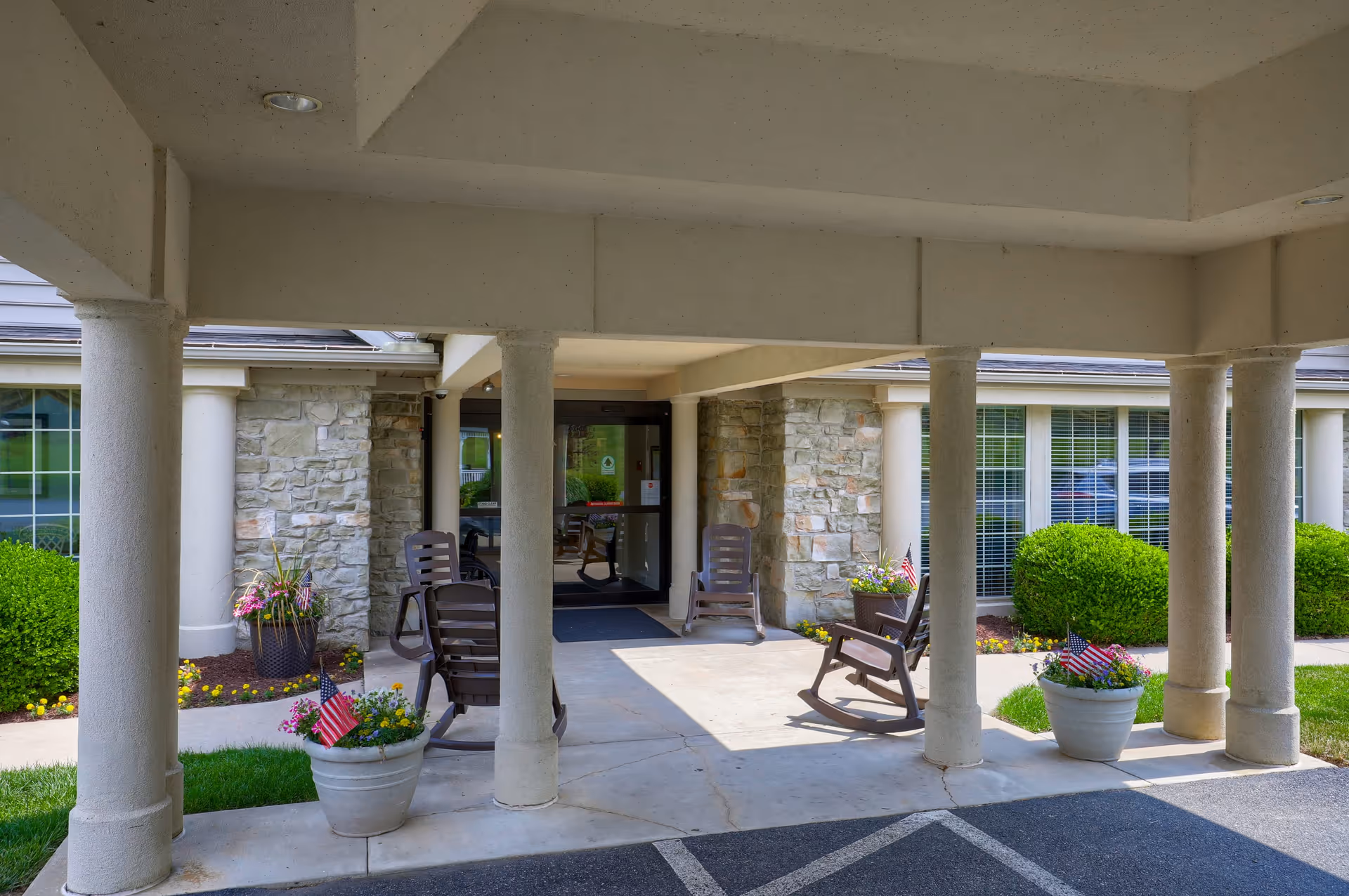 Covered entrance area of a senior living facility with stone walls, several brown rocking chairs, potted plants with small American flags, and large windows with white blinds.