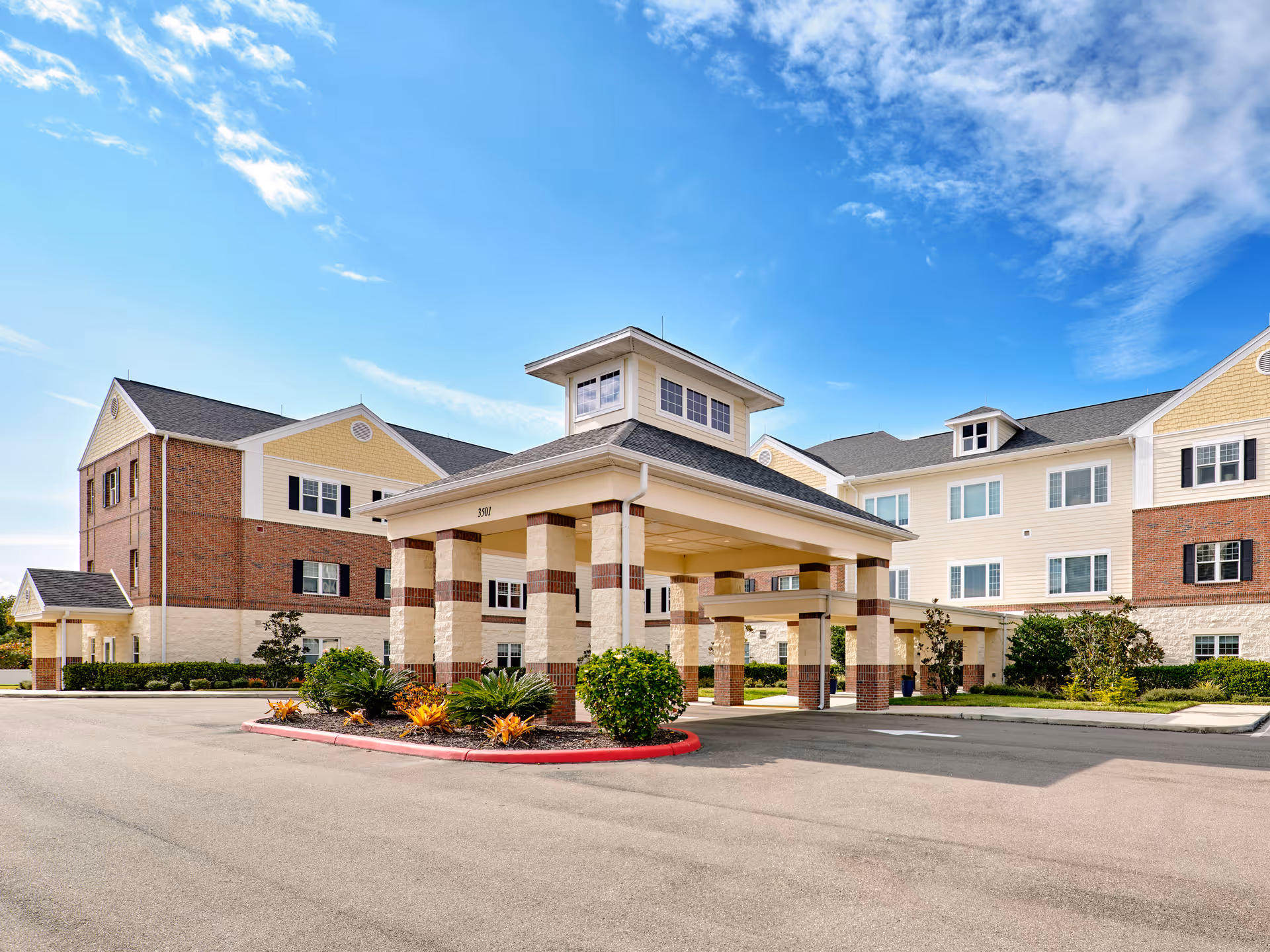 Senior living facility exterior with brick, beige siding, covered entrance, and landscaped circular driveway.