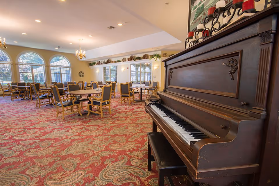 A spacious senior living facility common area with a red patterned carpet, multiple wooden tables and chairs, large arched windows letting in natural light, and a dark wooden upright piano in the foreground on the right side.