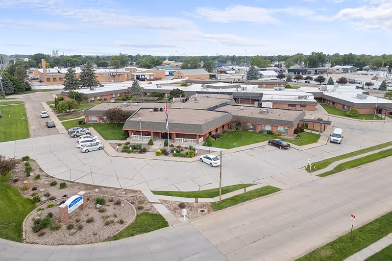 Aerial view of Good Samaritan Society - Hastings Village facility showing a large single-story building with multiple wings, surrounded by parking areas, green lawns, and landscaping. Several cars are parked near the building, and an American flag is visible in front of the main entrance.
