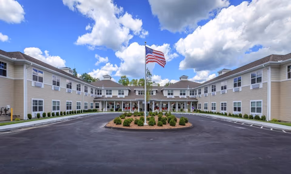 Front exterior view of a two-story assisted living facility building with beige siding and white trim. The building is U-shaped with a circular driveway in front, featuring a landscaped island with bushes and an American flag on a flagpole. The sky is blue with scattered clouds.
