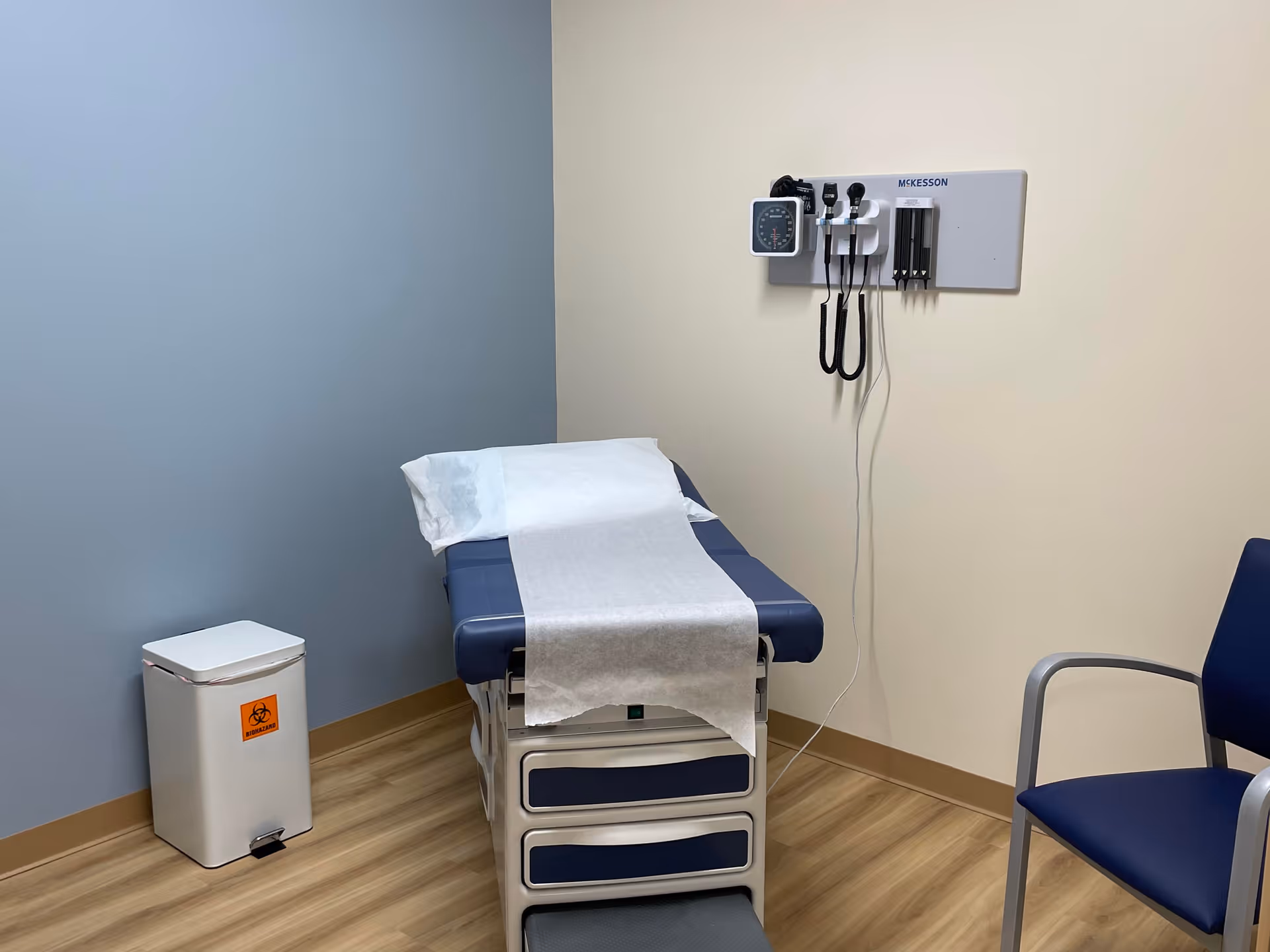 Medical examination room with wall-mounted monitor, exam table, trash can, and blue chair.