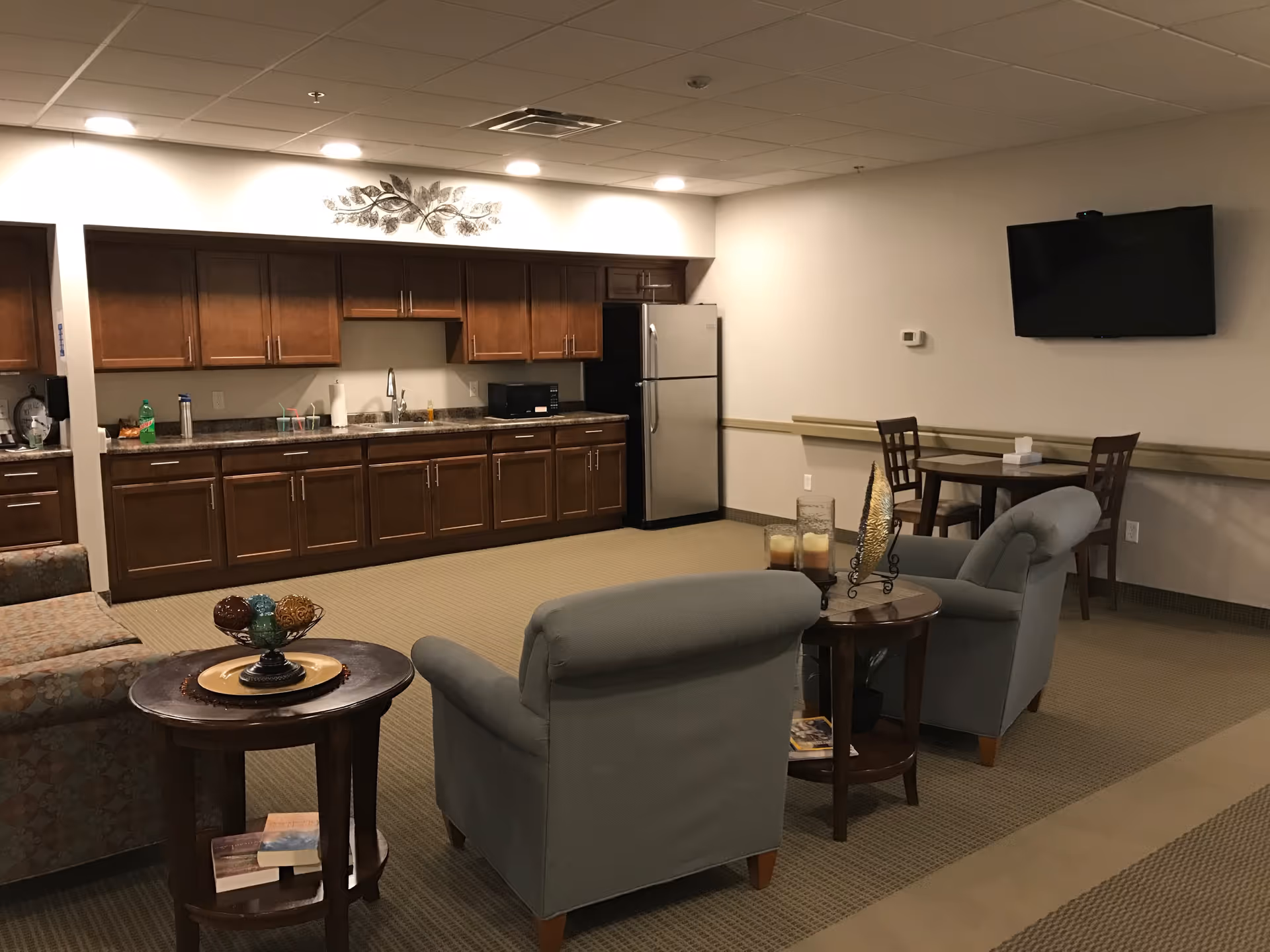 A communal lounge area with armchairs and side tables facing a wall-mounted TV, and a kitchenette with wooden cabinets and a refrigerator in the background.