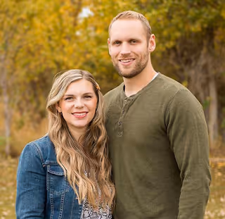 A couple stands outdoors with autumn trees in the background.