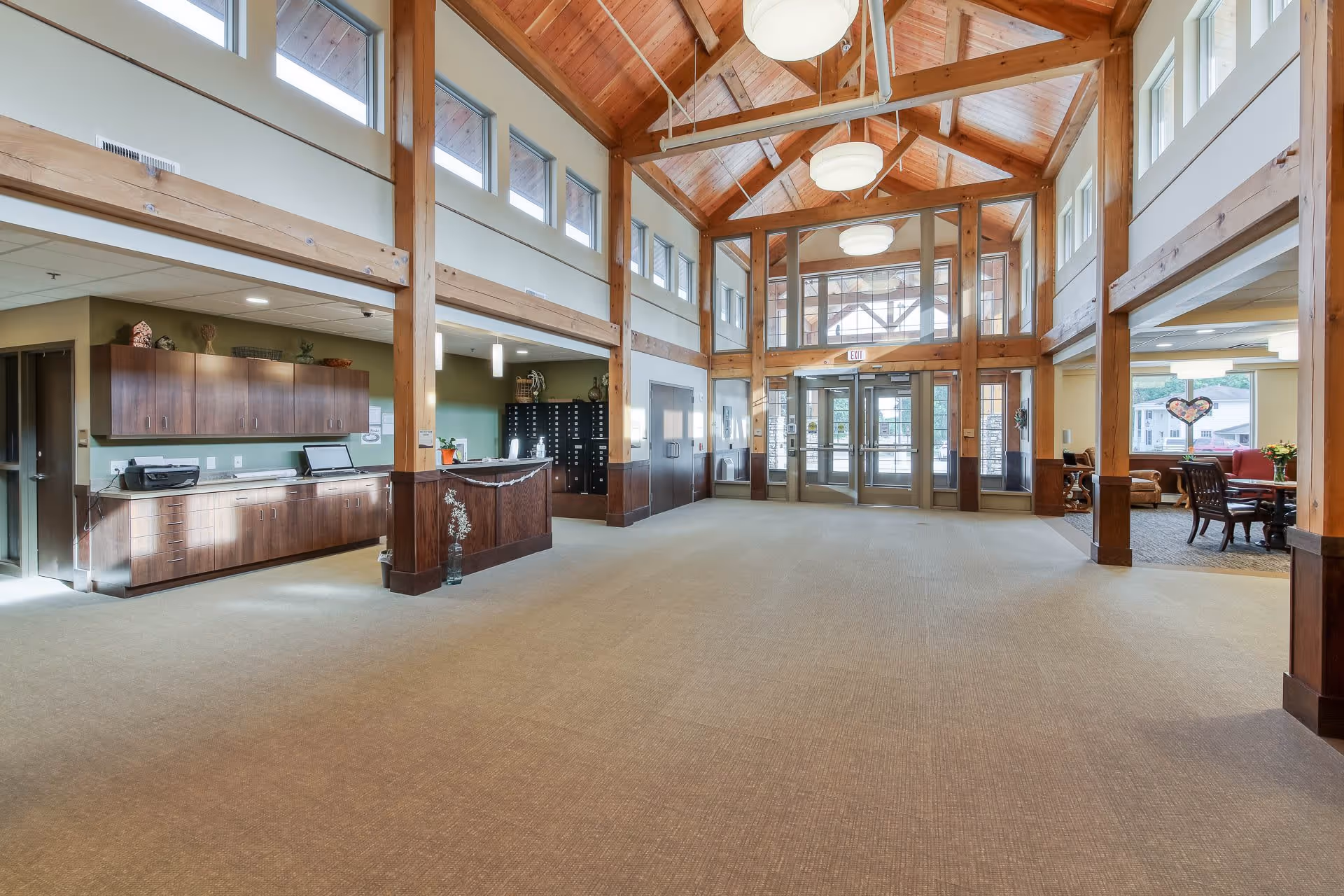 Spacious senior living facility lobby with high wooden beam ceilings, large windows letting in natural light, a reception desk on the left, and a seating area with chairs and tables on the right near large windows.