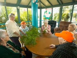 A group of elderly residents sit around a round table in a sunlit common room with large windows and plants.