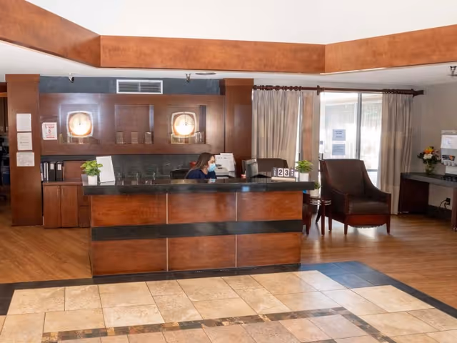 Reception area of Grossmont Post Acute Care facility with a wooden front desk, a staff member wearing a face mask seated behind the desk, two round wall lights, potted plants on the desk, a large armchair, and a window with curtains in the background.