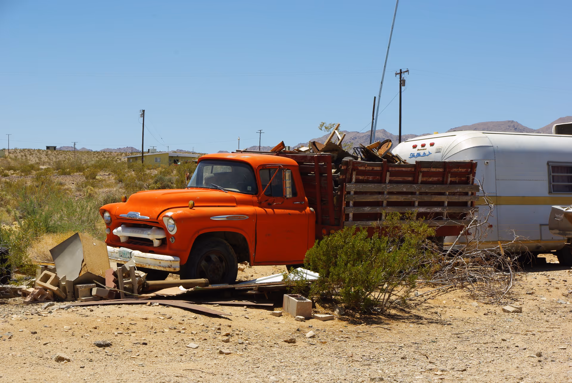 Old orange truck and wooden trailer clutter a desert junkyard with debris.