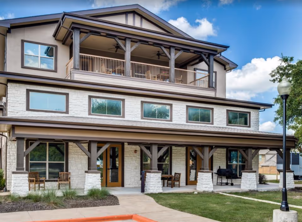 Exterior view of a two-story building with a covered porch supported by stone and wooden pillars. The building has large windows, outdoor seating with chairs and tables, a barbecue grill, and a lamp post on the lawn. The sky is blue with some clouds.