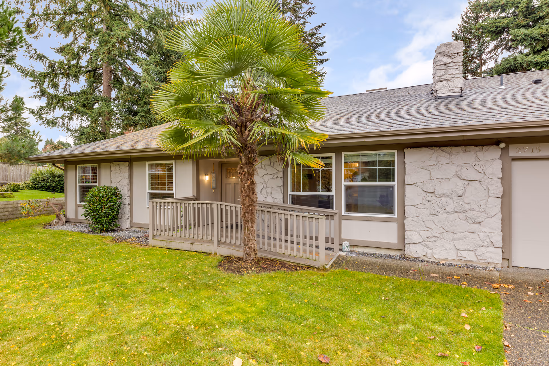 Single-story beige house with stone accents, wooden porch railing, palm tree, and green lawn.