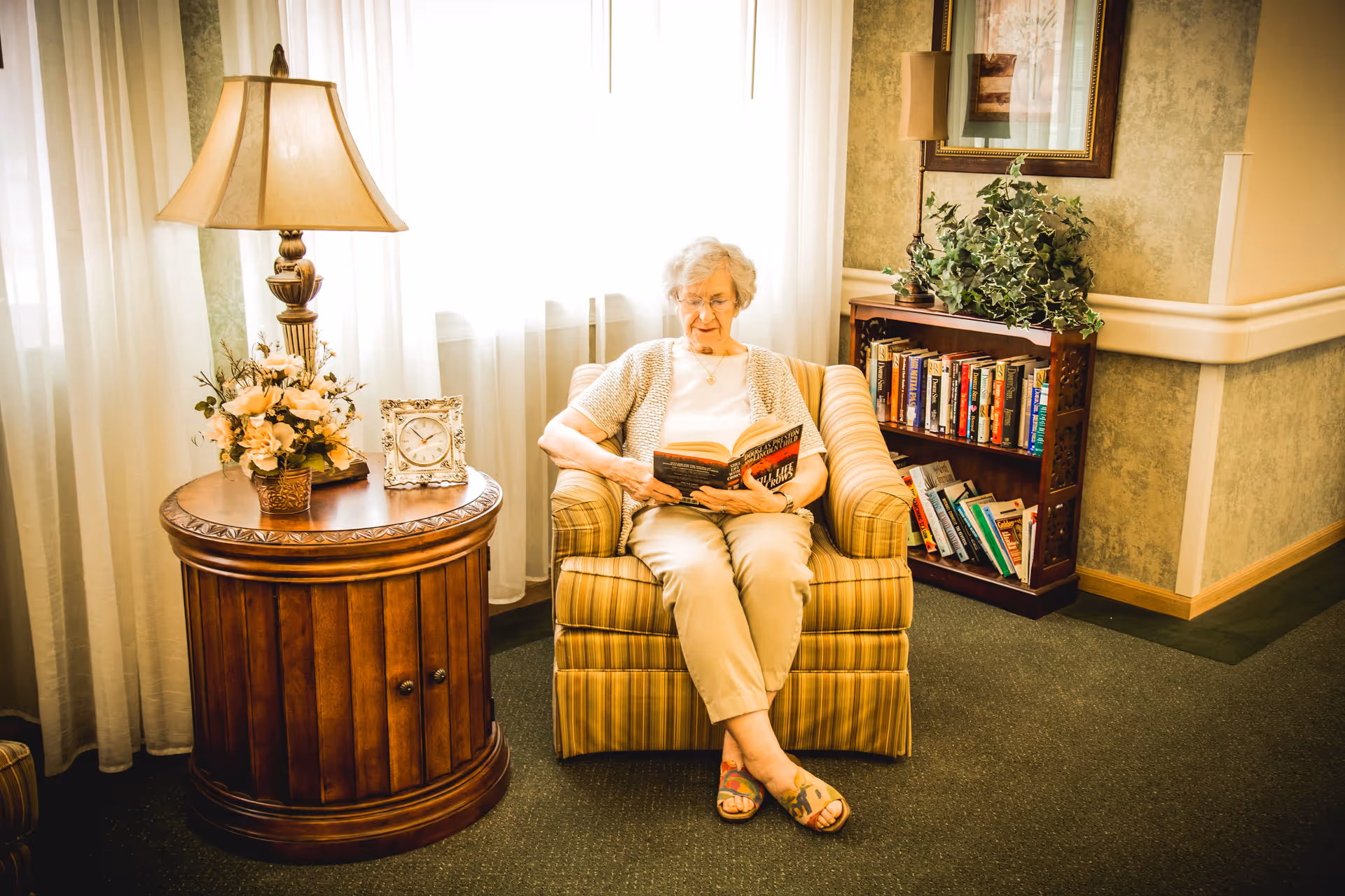 An elderly woman sitting in a striped armchair reading a book in a cozy room with a wooden side table holding a lamp, a flower arrangement, and a clock. Behind her is a bookshelf filled with books and a potted plant on top. The room has light-colored curtains and wallpaper with a framed picture on the wall.
