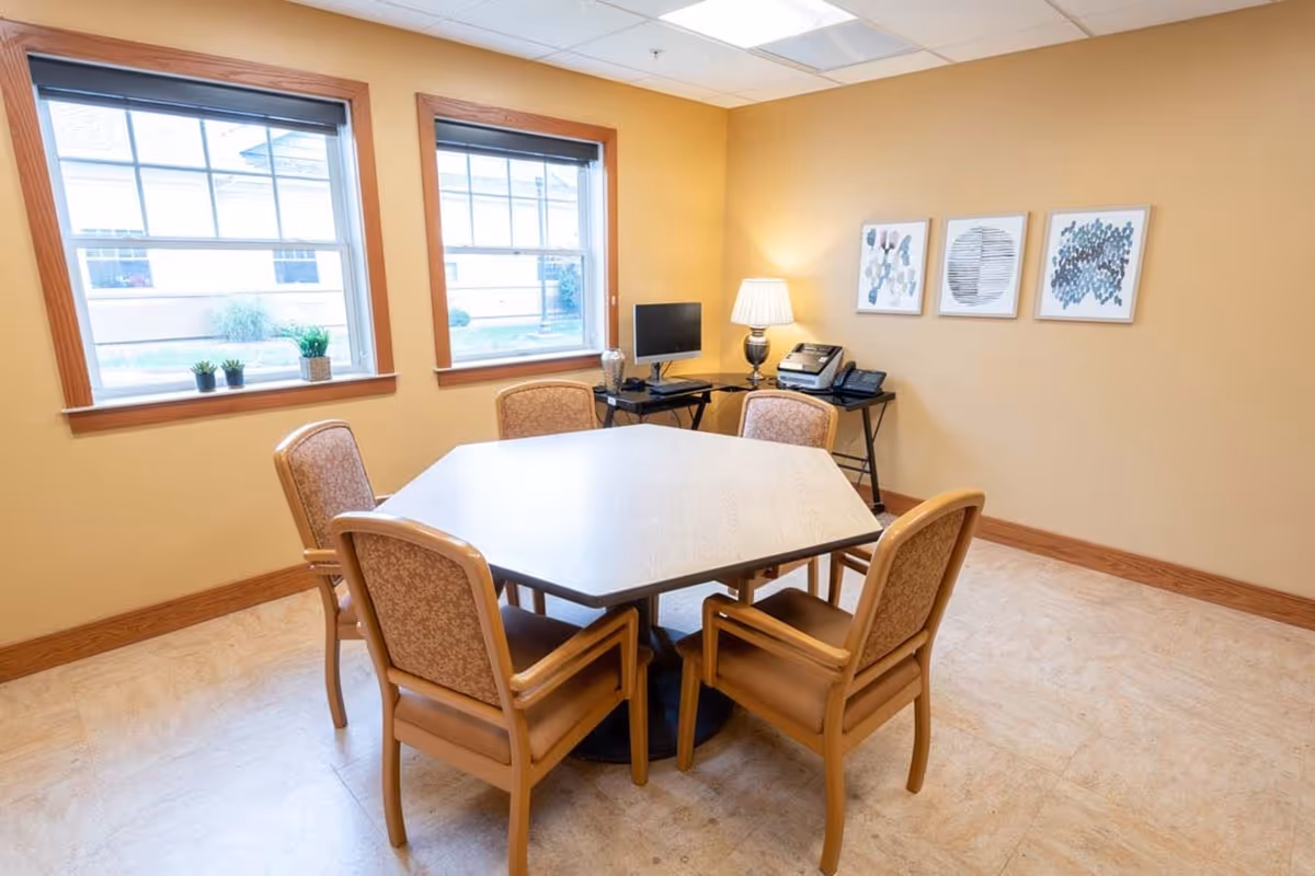A small meeting room with a square table and five beige chairs beside windows.