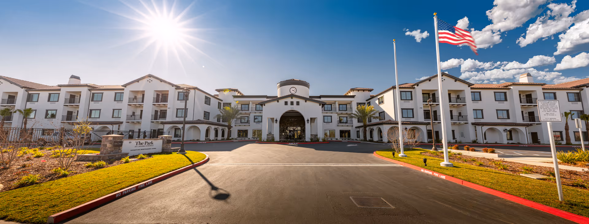 Sunlit exterior view of The Park at Laguna Springs with a circular driveway and flagpoles.