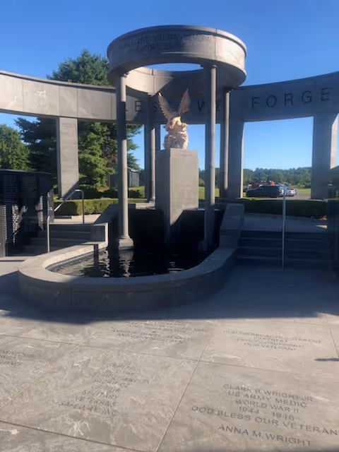 Outdoor memorial fountain with eagle statue and curved stone arches in a sunny plaza.