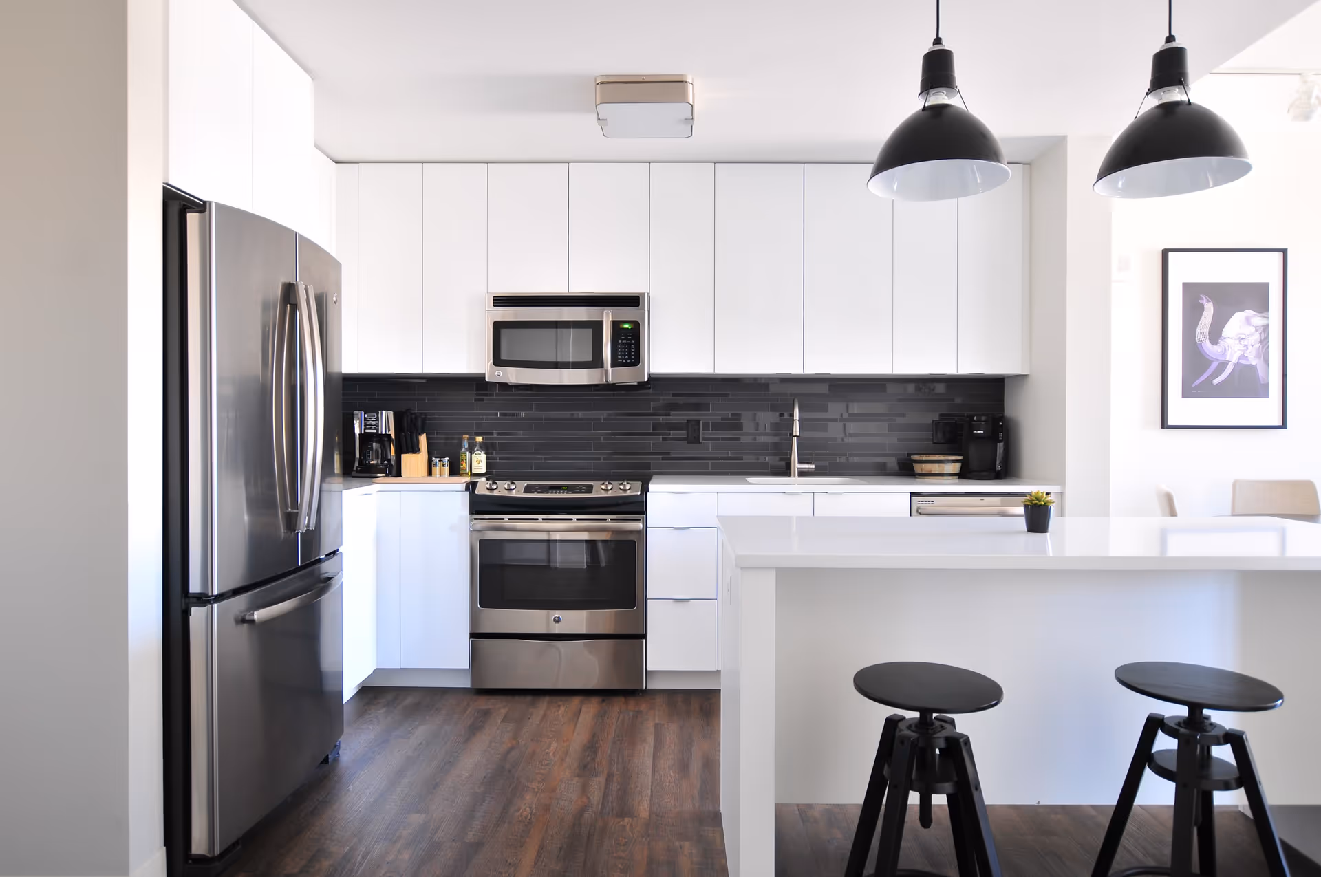 Modern kitchen with white cabinets, stainless steel refrigerator, oven, microwave, and dishwasher. The kitchen has a dark tile backsplash, wooden floor, a white island with two black stools, and two black pendant lights hanging from the ceiling. A framed artwork is visible on the wall to the right.