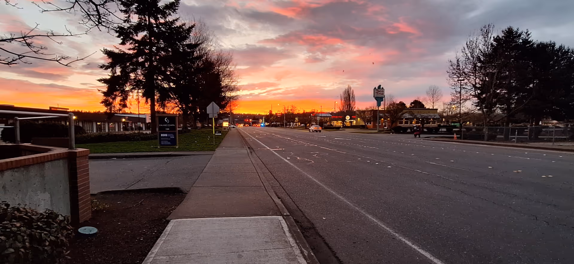 Sunset casts orange glow over a wide street, with sidewalk, trees, and distant buildings.