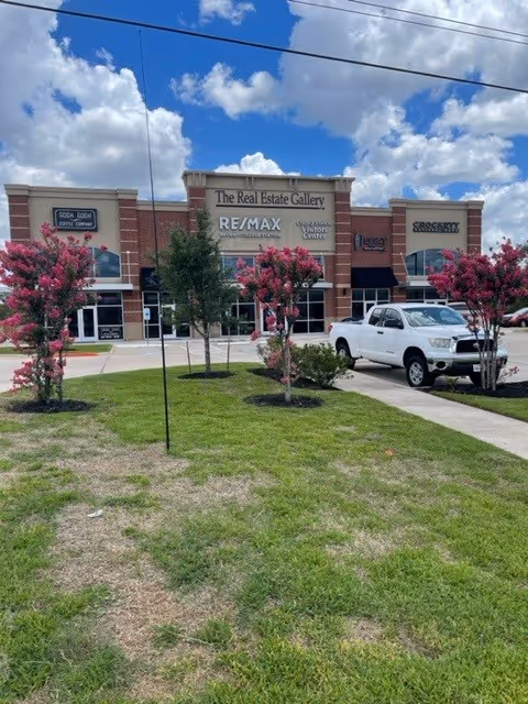Exterior of a strip mall with storefronts, pink flowering trees, and a white pickup.