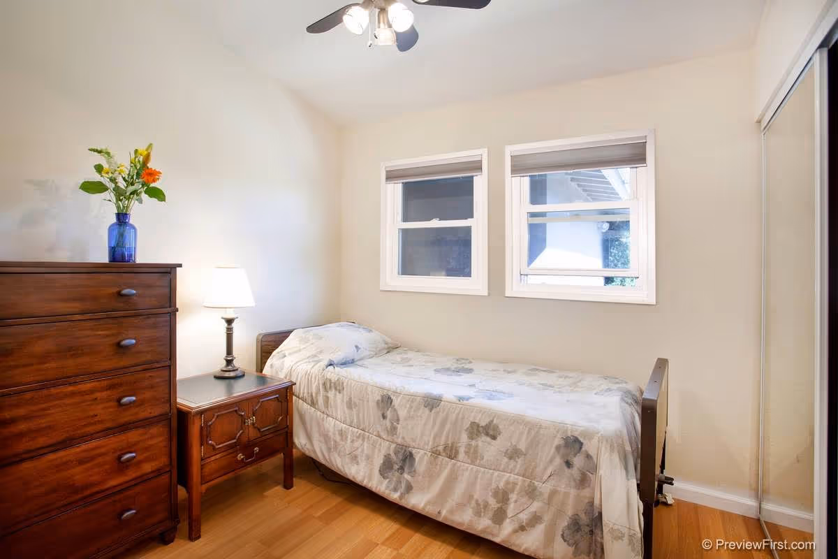 Small bedroom in a senior living facility with a single bed, dresser, and lamp.