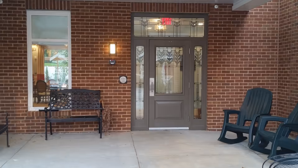 Covered entrance to a brick building with a decorative glass door, a bench, and plastic rocking chairs on the porch.