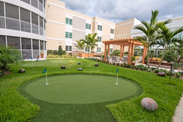 Courtyard featuring a small putting green with flags, surrounding lawn and landscaping, a wooden pergola with seating, and the multi-story building facade behind.