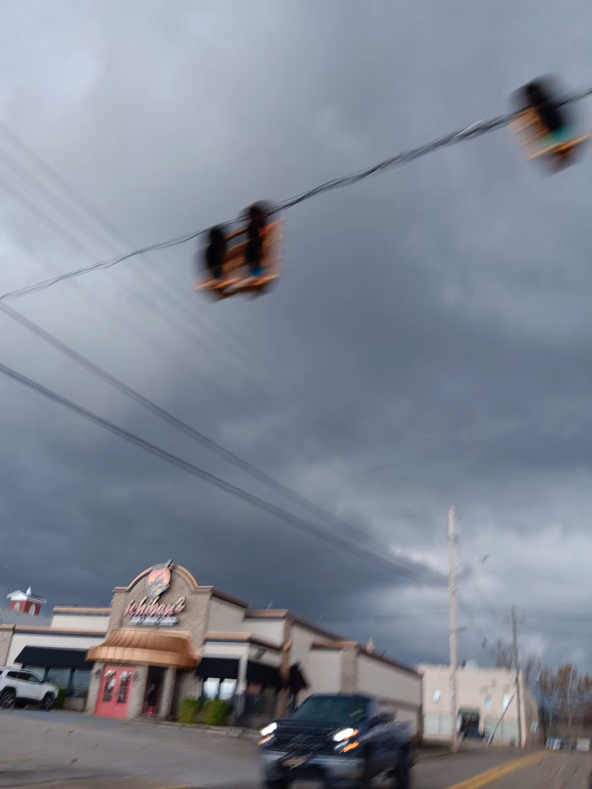 Blurry two people ride a suspended chair beneath a dark stormy sky near a storefront.
