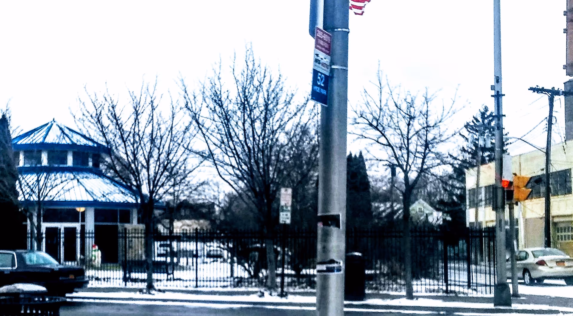 Fenced outdoor scene with bare trees, a blue-roofed pavilion, street lamps, and parked cars.