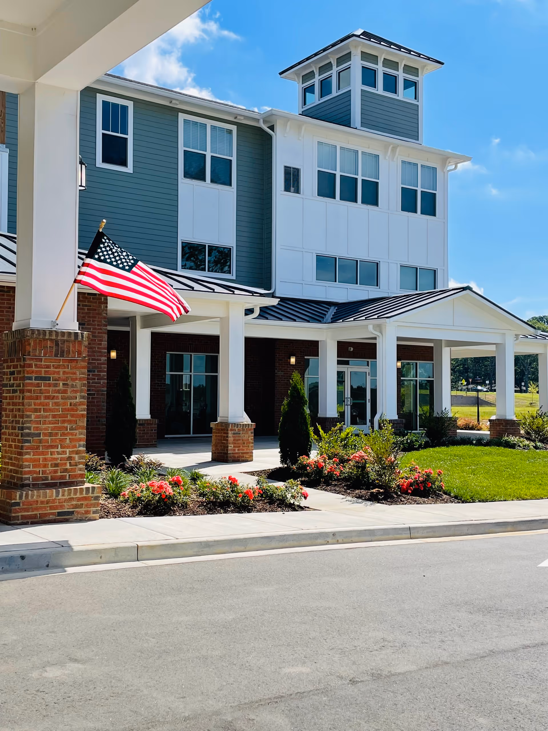 Front exterior view of a senior living facility building with a covered entrance supported by white columns and brick bases. There is an American flag mounted on one of the columns, flower beds with red flowers, green shrubs, and a well-maintained lawn. The building has multiple windows and a tower-like structure on the roof under a clear blue sky.