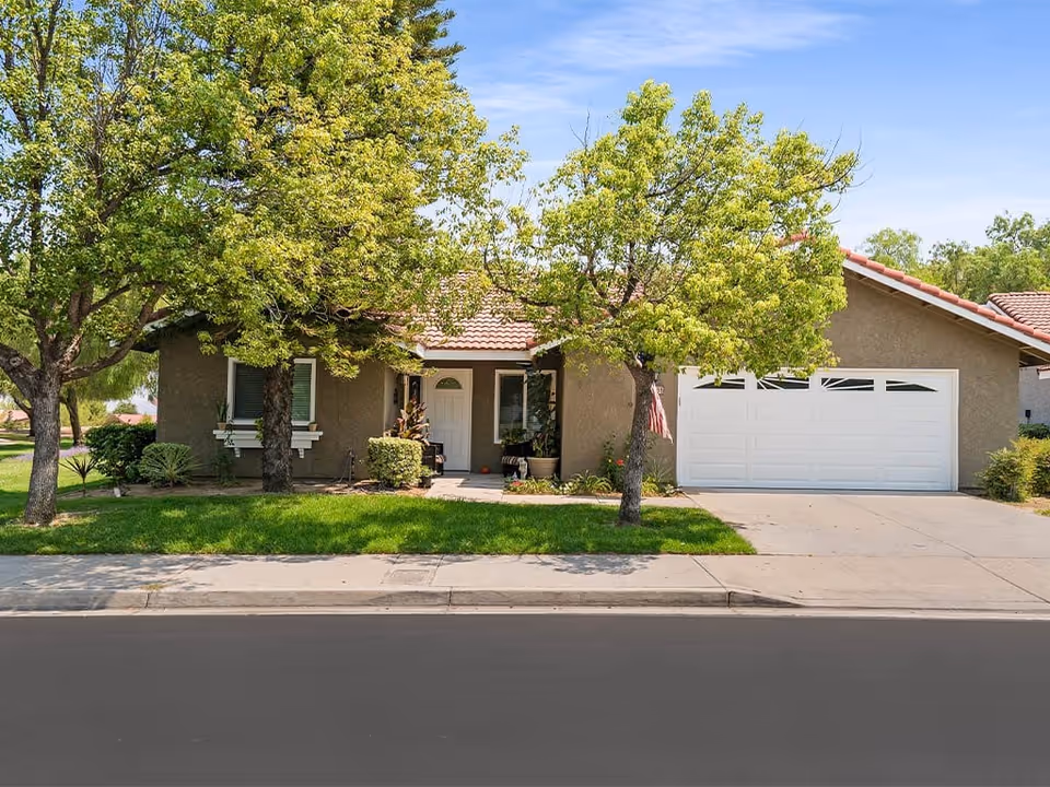 Single-story house with a two-car garage, front lawn and trees along the driveway under a blue sky.