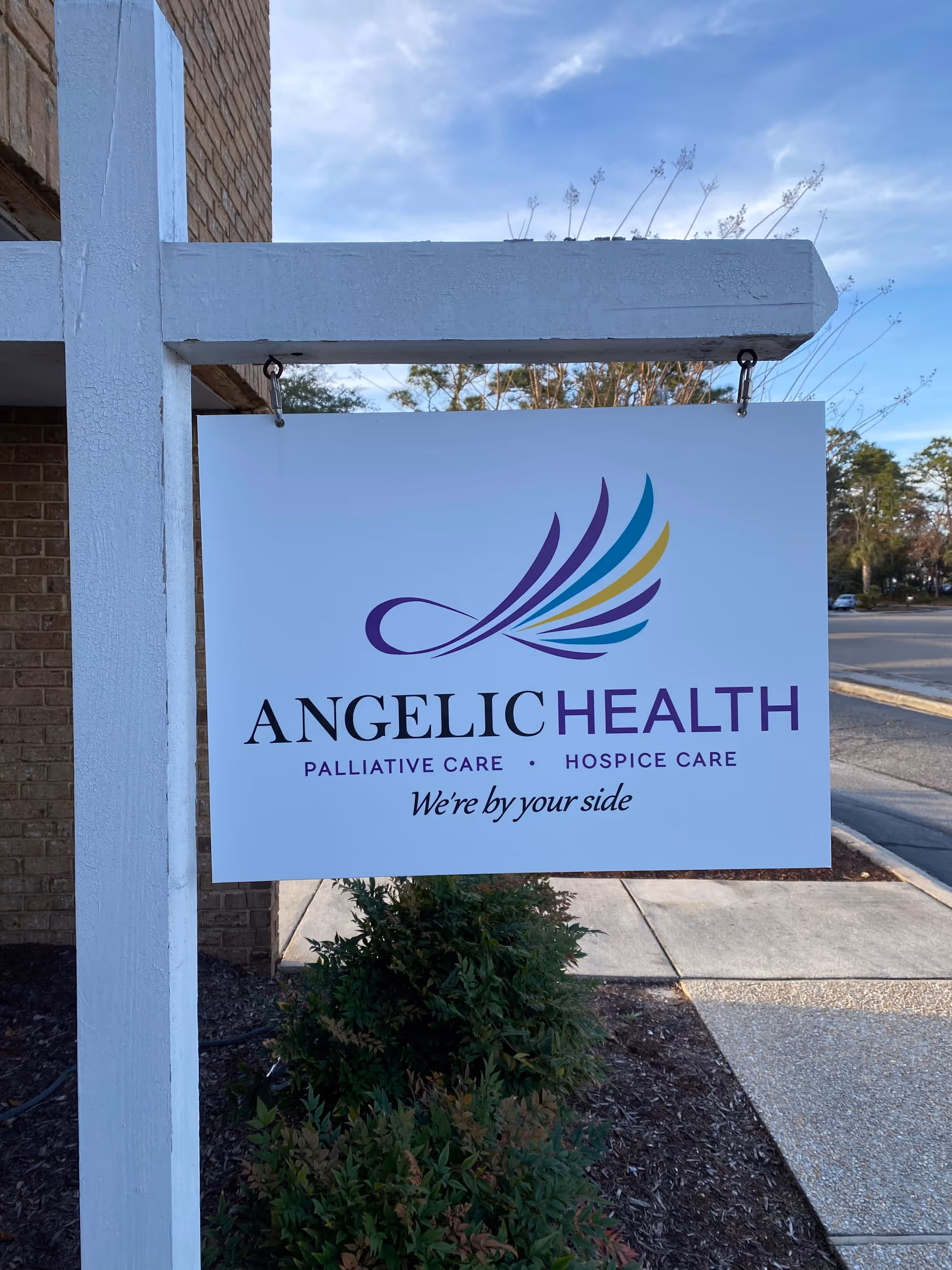 Angelic Health sign with colorful wings hangs on a white post beside a sidewalk.