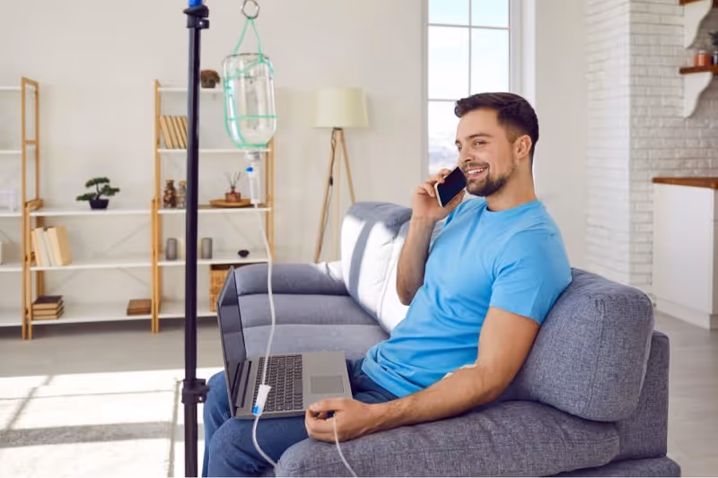 A man sits on a sofa in a living room with an IV drip and laptop.