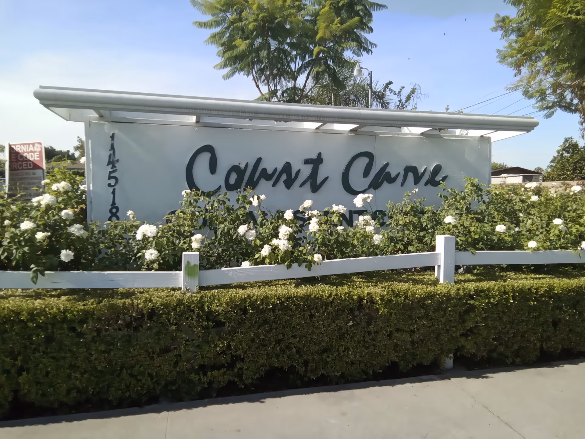 Entrance sign reading 'Coast Care' surrounded by white roses, a low hedge and a white fence.