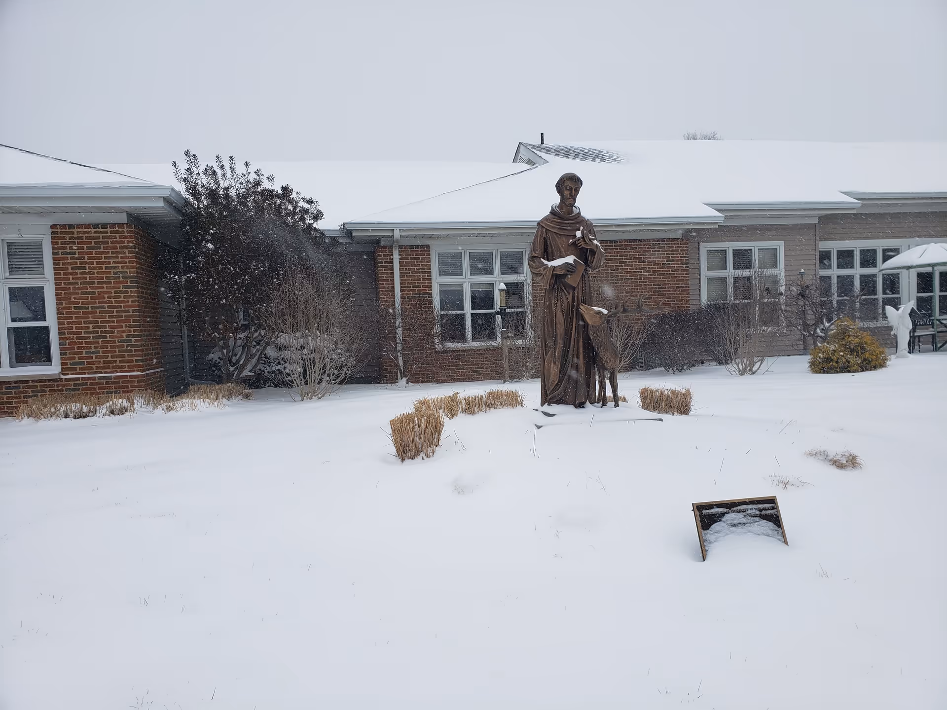 Snow blankets the front of a Mercy Hospice brick building with a statue.