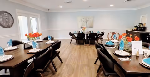 A bright dining room with wooden floors and light blue walls. There are two dark wooden dining tables set with white plates, blue napkins, glasses, and orange flower centerpieces. A large clock is on the left wall, and a painting hangs on the far wall. French doors are visible on the left side of the room.