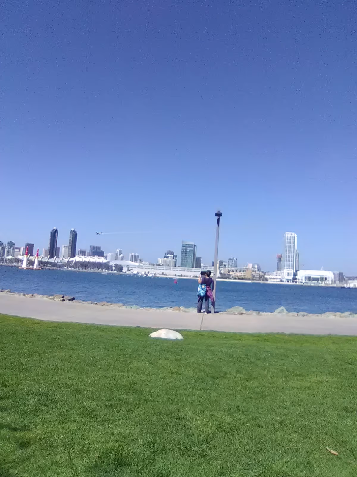Two people stand by a waterfront park with grass, water, and city skyline.