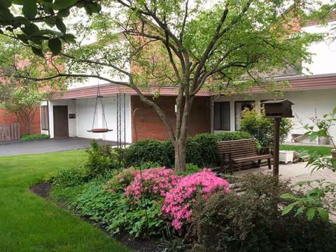 Outdoor garden area with a tree, flowering shrubs, and a bench near the facility entrance.