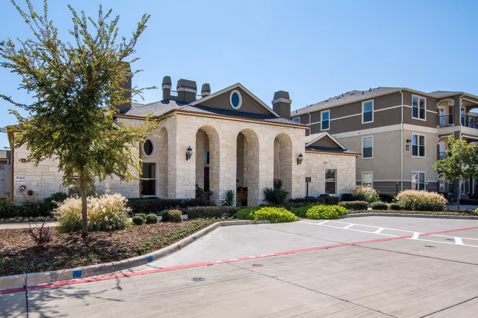 Front view of a stone-faced community building with three arched entrances, landscaped grounds, and adjacent multi-story residential buildings under a clear sky.