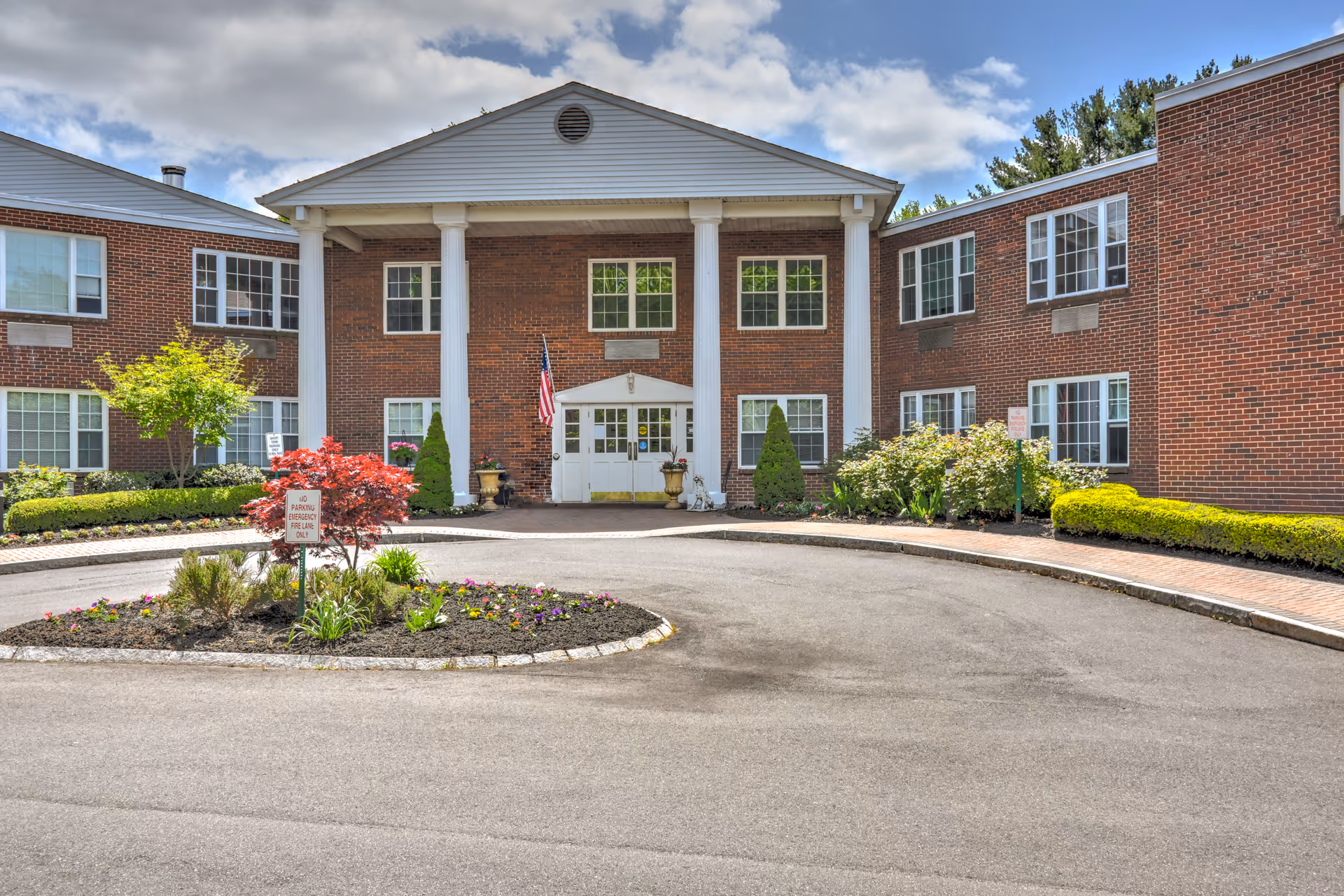 Brick senior living facility with white columns, flag, and landscaped circular driveway.