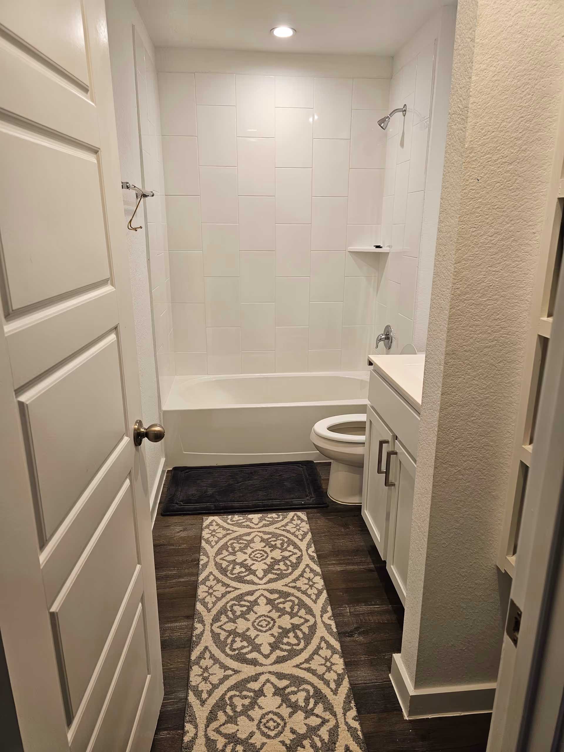 Small bathroom with a white tiled shower, toilet, vanity, and patterned rug.