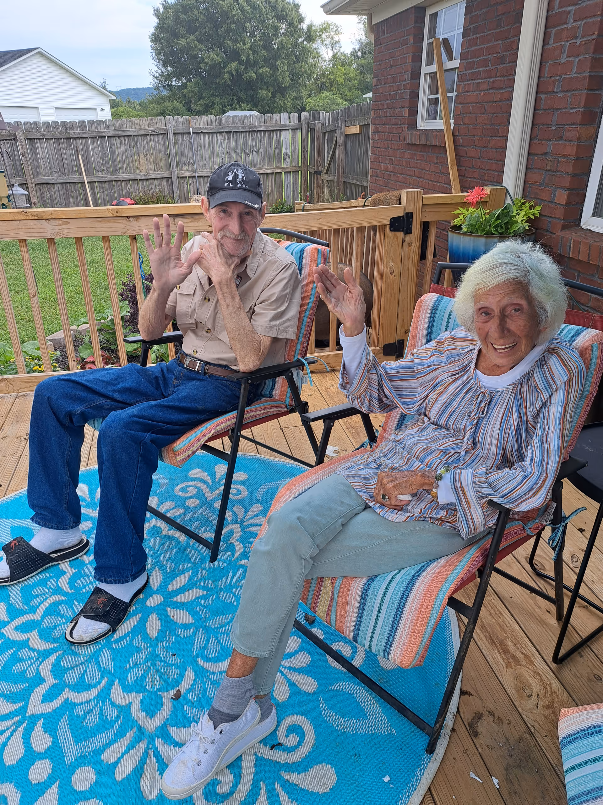 Two elderly residents sit on a wooden deck, waving, in striped chairs on an outdoor patio.