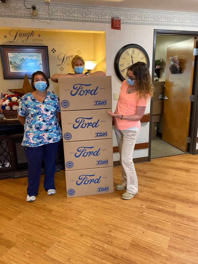 Three masked staff stand in a facility interior beside a tall stack of Ford-branded boxes.