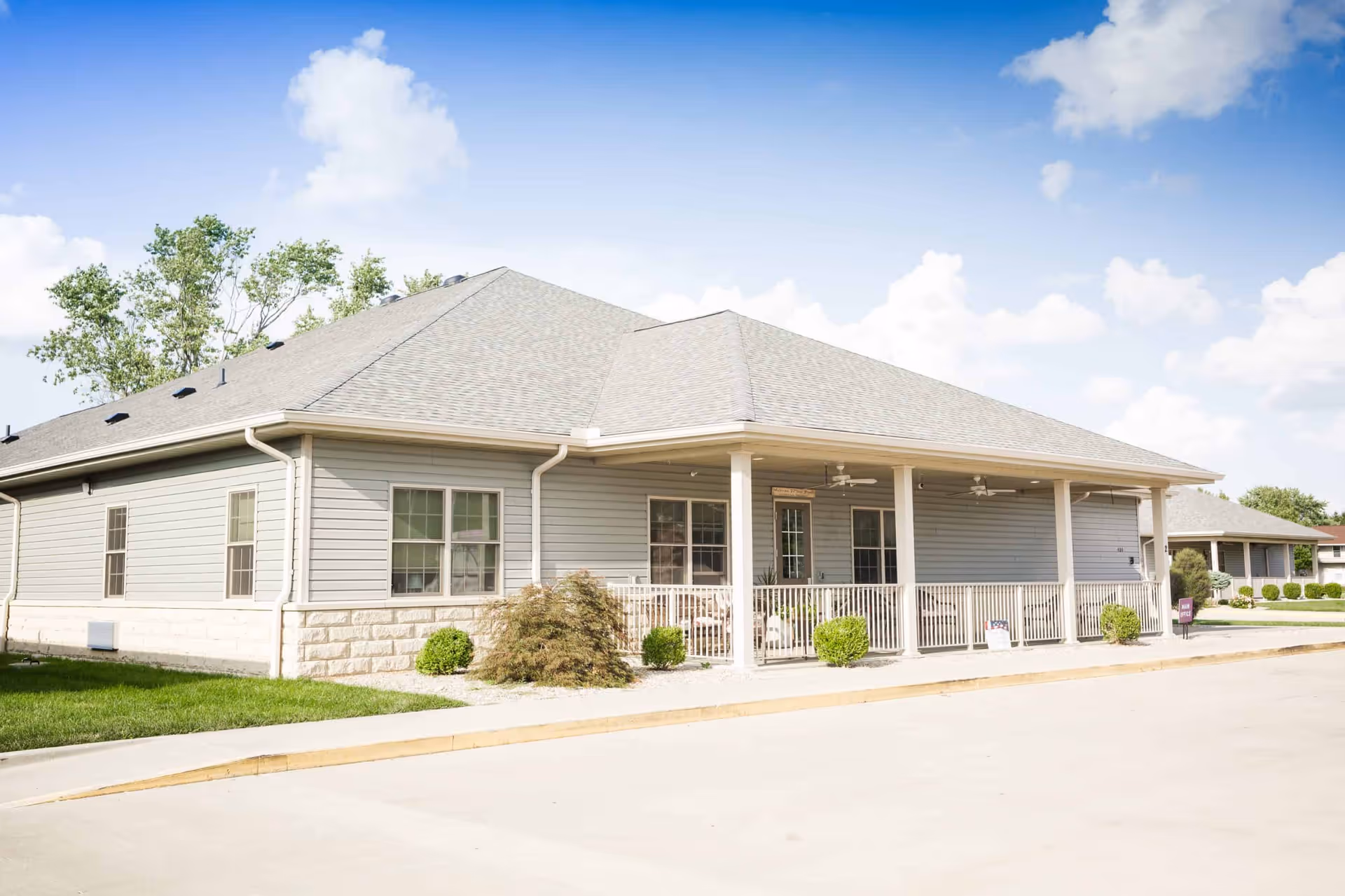 Exterior view of a single-story building with light gray siding and a covered porch with white railings. The building is surrounded by a well-maintained lawn and small bushes, under a blue sky with scattered clouds.
