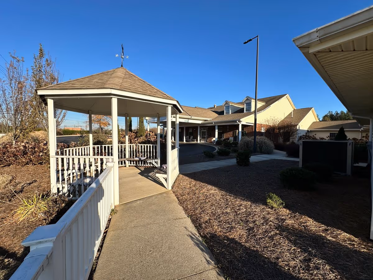 A white wooden gazebo and walkway leading to the front entrance of a single-story assisted living building under a clear blue sky.