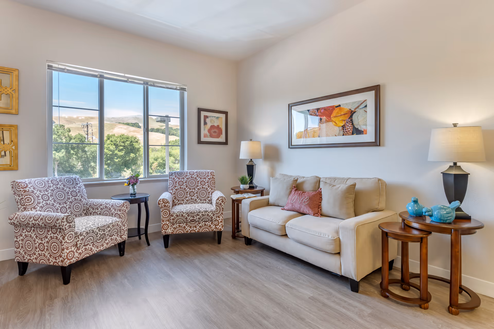 Sunlit sitting area with two patterned armchairs, a beige sofa, side tables, lamps, and a large window with a landscape view.