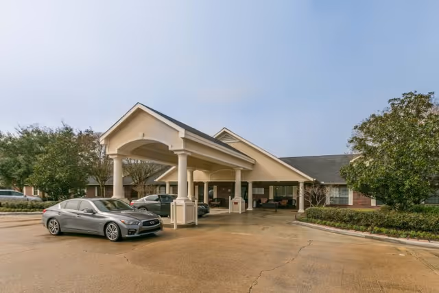 Exterior view of a single-story building with a covered entrance supported by columns. Several cars are parked in front of the building, and there are trees and shrubs around the parking area under a clear sky.
