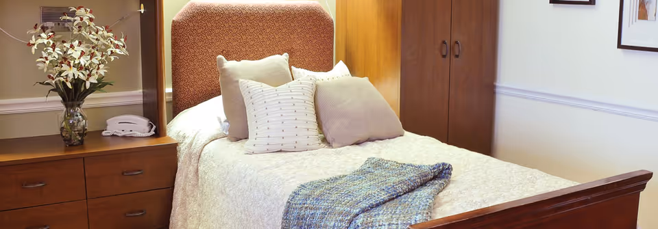 A neatly made single bed with pillows and a folded blanket in a cozy bedroom featuring a wooden dresser, wardrobe, and a vase of flowers on the nightstand.